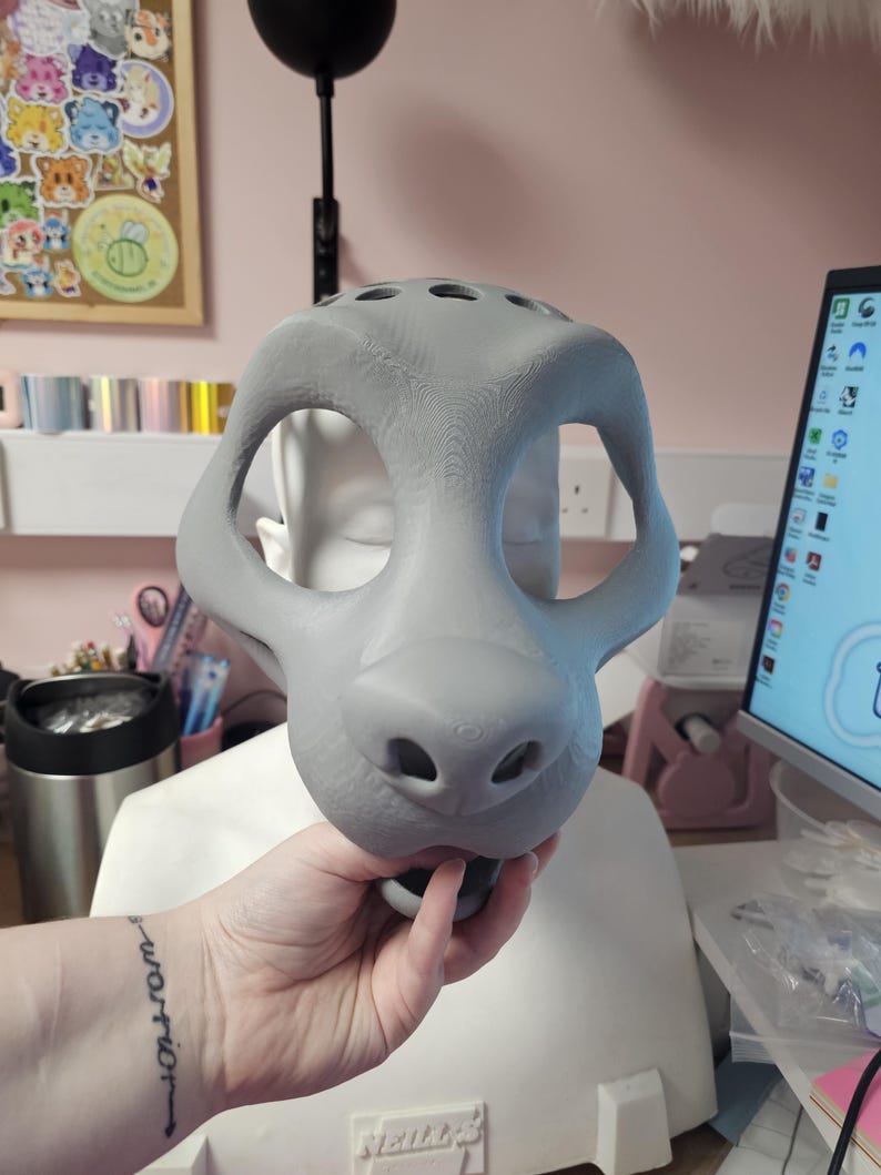 May include: A gray, unfinished fursuit head base, held by a hand. The base has large eye openings, a prominent snout with nostrils, and several ventilation holes on top. The overall shape suggests a canine or feline design.