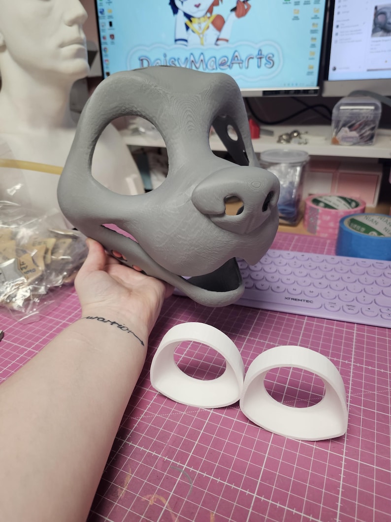 May include: A gray, 3D-printed animal mask with a snout and eye openings, held by a person. Two white, curved eye pieces are in front of the mask. The mask is likely for cosplay or costume use.