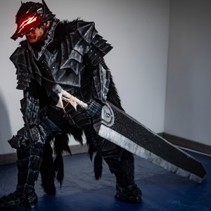 Puede incluir: Una persona con un traje negro y plateado con una espada grande. El traje es detallado e incluye un casco con ojos rojos brillantes.