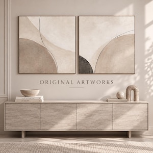 Pode incluir: Duas pinturas quadradas em tela com formas curvas abstratas em bege, creme e cinza escuro com linhas pretas finas. Abaixo da arte, um aparador longo de pedra clara contém uma tigela, dois livros empilhados, uma esfera e uma escultura em arco.