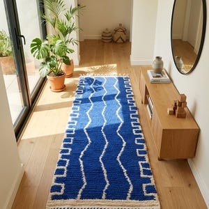 May include: A blue and white patterned runner rug with a zig-zag design and fringe. The rug is placed on a hardwood floor in a hallway. The rug's design features a repeating pattern of white lines against a blue background.