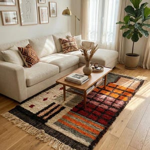 May include: A living room scene featuring a cream-colored sectional sofa with decorative throw pillows. A wooden coffee table holds books and a vase with dried flowers. A colorful rug with geometric patterns and fringe adds warmth to the space.