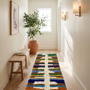 Puede incluir: Una alfombra de pasillo con un patrón geométrico en tonos azules, marrones, verdes y blancos. La alfombra tiene un borde con flecos y está colocada sobre un suelo de madera. También se ven un banco de madera y una planta en maceta.