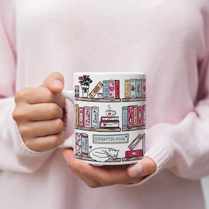May include: White ceramic mug with a colorful illustration of a bookshelf filled with books, a cat, and a cup of tea. The text "STFWTTDLAGG" is printed on the shelf.