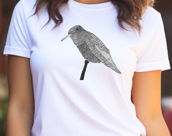 May include: A white t-shirt with a detailed black and white bird illustration. The bird is perched on a black stem. The person in the image is wearing a white hat and pink trousers. The background is blurred, suggesting an outdoor setting.