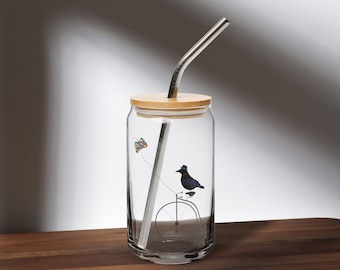 May include: A clear glass drinking tumbler with a bamboo lid and a stainless steel straw. The tumbler has a colorful butterfly and a black bird riding a bicycle printed on the side.