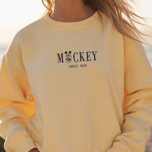 May include: A pale yellow sweatshirt with the word "MOCKEY" and a Mickey Mouse graphic, along with "SINCE 1928" printed in black. The word "BUTTER" is printed in a stylized font at the top left. The sweatshirt has long sleeves and a crew neck.