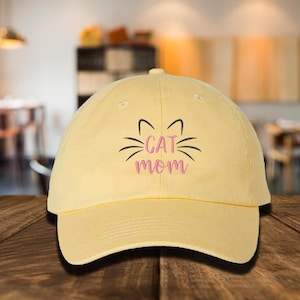 May include: A yellow baseball cap with a pink embroidered design that says "CAT MOM" with a cat face above the text.