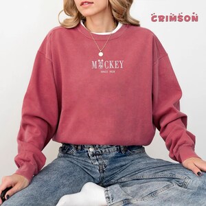 May include: A crimson-colored sweatshirt with the word "Mickey" and a Mickey Mouse graphic in white, along with "Since 1928." The sweatshirt has a crew neck and long sleeves. The word "Crimson" is written in a bubble font in the top right corner.