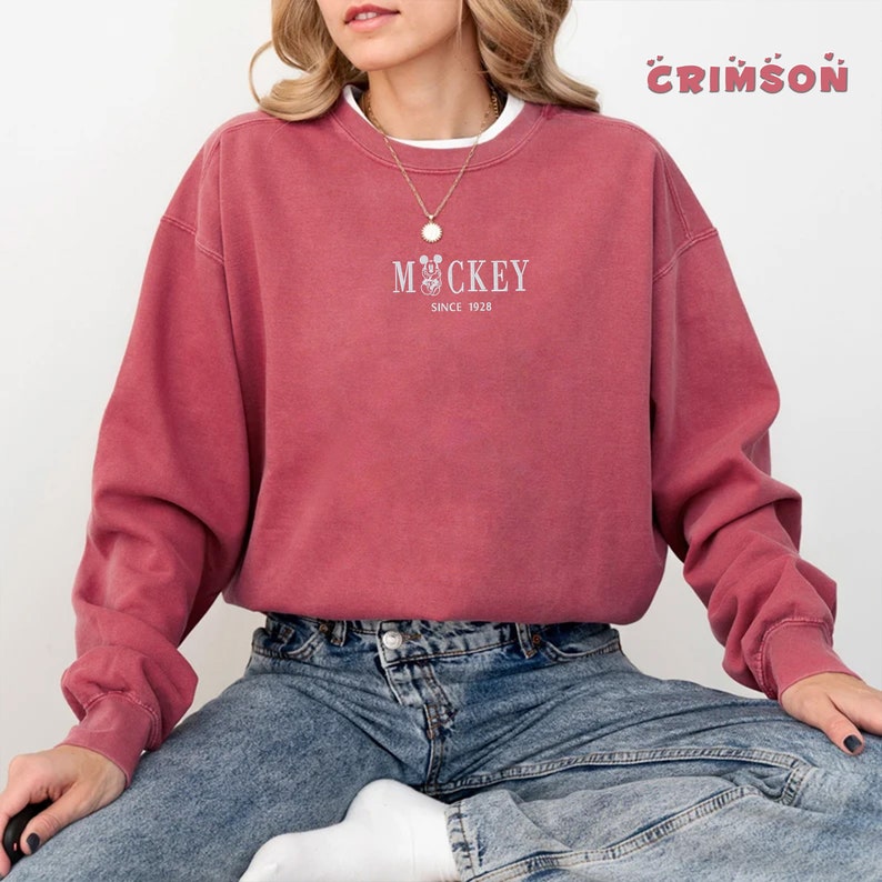 May include: A crimson-colored sweatshirt with the word "Mickey" and a Mickey Mouse graphic in white, along with "Since 1928." The sweatshirt has a crew neck and long sleeves. The word "Crimson" is written in a bubble font in the top right corner.