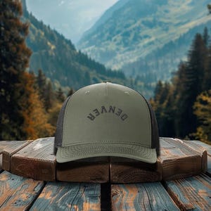 May include: An olive green trucker hat with the word "DENVER" arched across the front. The hat has a black mesh back and a curved brim. The hat is sitting on a wooden surface with a mountain backdrop.