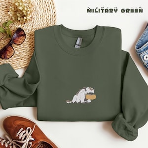 May include: A military green crewneck sweatshirt featuring a small embroidered graphic of a white and brown creature holding a tan object. The text "MILITARY GREEN" is printed in the upper right corner of the garment in a dark font.