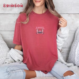 Puede incluir: Camiseta de color rojo carmes&iacute; con un peque&ntilde;o dise&ntilde;o bordado de un personaje de dibujos animados rosa con la palabra "ANGEL" encima. La camiseta es de color liso y tiene cuello redondo.