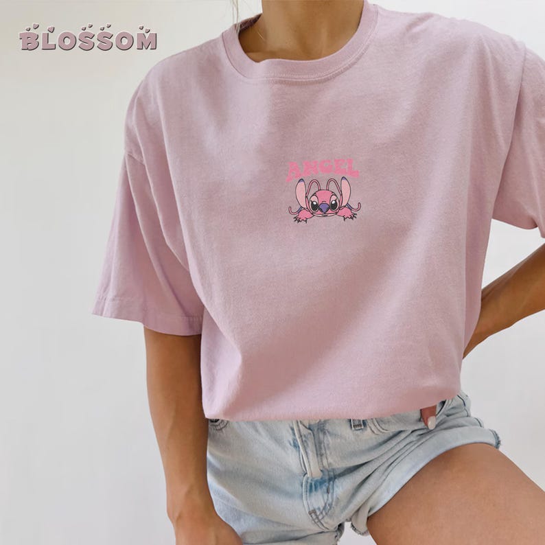 Puede incluir: Camiseta rosa claro con un personaje de dibujos animados y la palabra "ANGEL" encima. La camiseta tiene mangas cortas y un corte holgado. La palabra "BLOSSOM" est&aacute; impresa en la esquina superior izquierda.