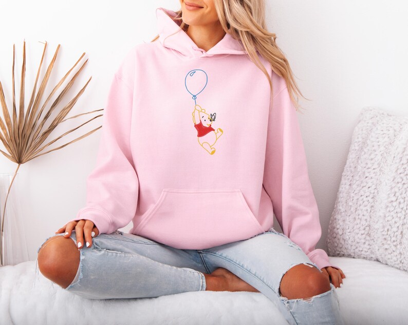 Puede incluir: Una sudadera con capucha rosa claro con un bolsillo de canguro y una capucha con cord&oacute;n. La parte delantera presenta una imagen bordada de un oso de dibujos animados agarrando un globo azul. El oso lleva una camisa roja.