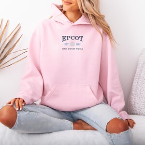 May include: A light pink hoodie with a front pocket and a hood. The text "EPCOT EST. 1982 WALT DISNEY WORLD" is printed in navy blue on the chest. The hoodie is made of a soft material.