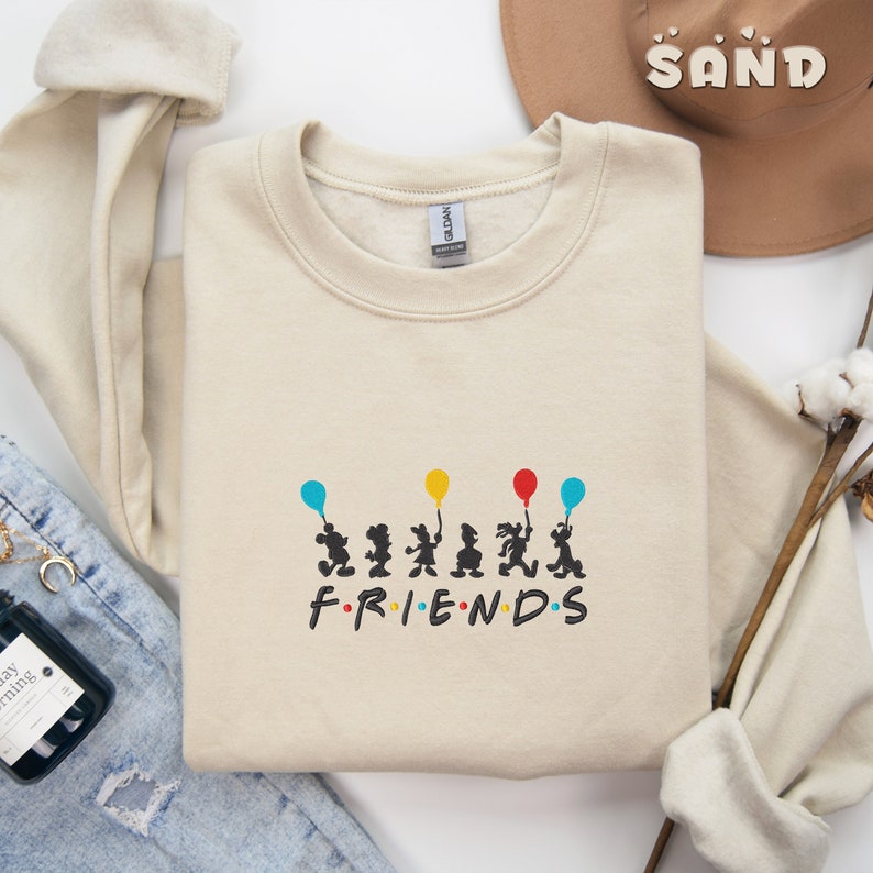 May include: A beige sweatshirt with a graphic of six cartoon figures holding balloons and the word "FRIENDS" printed below. The figures are black silhouettes with colorful balloons.