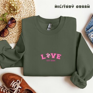May include: A military green sweatshirt featuring the pink embroidered word "LOVE" and the text "EST 2026". The crew neck sweatshirt is made of a soft material. The image also includes sunglasses and a woven basket.