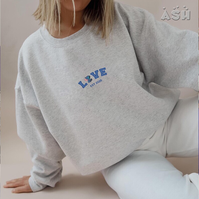 May include: A light grey crew neck sweatshirt. The word "LOVE" is embroidered in blue, with a small floral design and the text "EST 2026" below. The sweatshirt is a casual style.