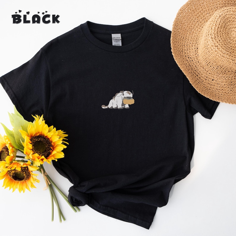 May include: A black short-sleeved t-shirt featuring a small embroidered graphic of a white and grey creature with horns holding a brown object. The shirt has a crew neckline and is displayed alongside a straw hat and a bouquet of yellow sunflowers.