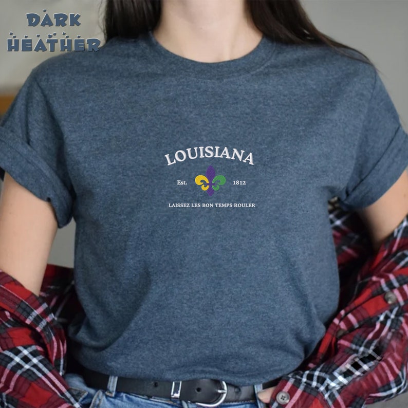 May include: A dark heather grey t-shirt with a white graphic design featuring the text "LOUISIANA" with a fleur-de-lis symbol and the text "Est. 1812" and "LAISSEZ LES BON TEMPS ROULER".