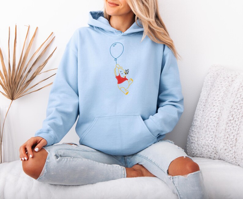 Puede incluir: Sudadera con capucha azul claro con bolsillo de canguro y capucha con cord&oacute;n. La parte delantera presenta un dise&ntilde;o bordado de un oso de dibujos animados agarrando un globo azul. El oso lleva una camisa roja y pantalones cortos amarillos.