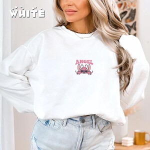 Pode incluir: Camisola branca com a palavra "WHITE" impressa no canto superior esquerdo e um gr&aacute;fico rosa de uma personagem de desenho animado com a palavra "ANGEL" por cima. A camisola de manga comprida &eacute; feita de um material macio.