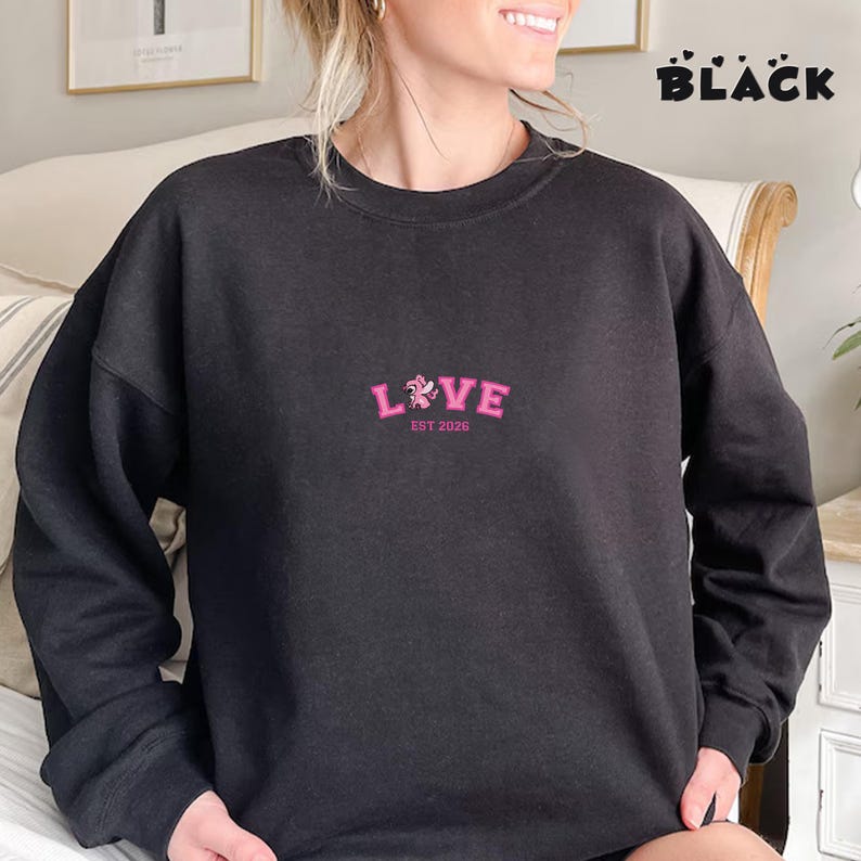 May include: Black sweatshirt featuring the word "LOVE" embroidered in pink, with a floral design and the text "EST 2026". The word "BLACK" is printed in a bold font at the top right corner of the image.