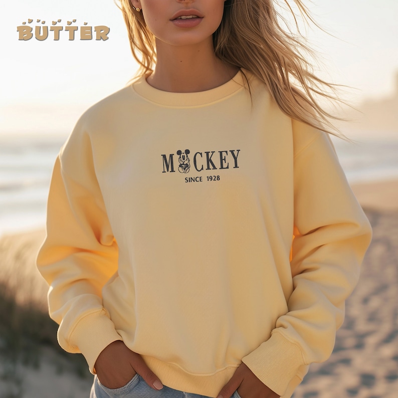 May include: A pale yellow sweatshirt with the word "MOCKEY" and a Mickey Mouse graphic, along with "SINCE 1928" printed in black. The word "BUTTER" is printed in a stylized font at the top left. The sweatshirt has long sleeves and a crew neck.