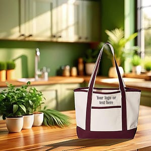 May include: A white tote bag with claret-coloured accents, including the base and handles. The bag features a pocket with the text "Your logo or text here". The bag is on a wooden surface.