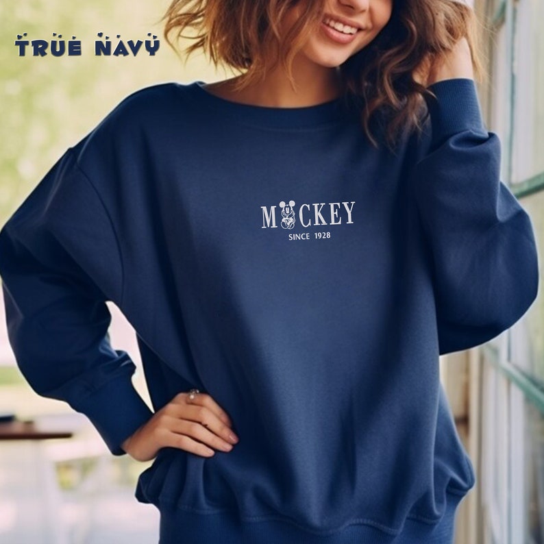 May include: A navy blue sweatshirt with the text "Mickey Since 1928" in white. The word "Mickey" is above a graphic of Mickey Mouse. The sweatshirt has long sleeves and a crew neck. The words "True Navy" are at the top left.