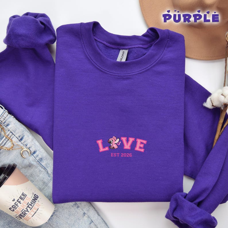 May include: A vibrant purple sweatshirt featuring the embroidered word "LOVE" in pink, accompanied by a floral motif and the text "EST 2026". The garment is folded, with glimpses of blue jeans and a coffee cup in the background.