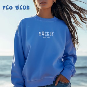 May include: A light blue sweatshirt with the text "FLO BLUE" at the top and "Mickey Since 1928" in the center. The sweatshirt is a crew neck style and is being worn by a person.