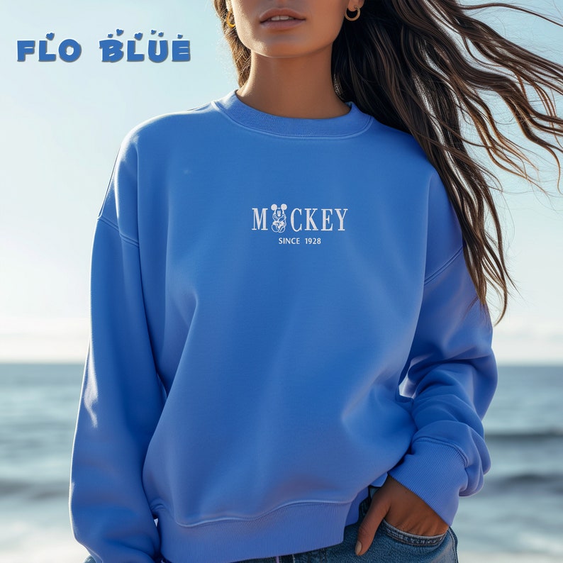 May include: A light blue sweatshirt with the text "FLO BLUE" at the top and "Mickey Since 1928" in the center. The sweatshirt is a crew neck style and is being worn by a person.