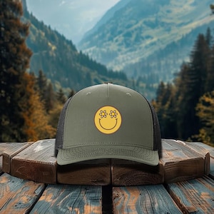 May include: Olive green trucker hat with a black mesh back. The hat features a yellow smiley face with daisy eyes. The hat is sitting on a wooden surface with a mountain backdrop.
