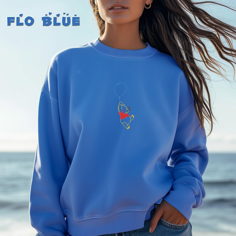 May include: A blue sweatshirt with a Winnie the Pooh graphic. The graphic shows Winnie the Pooh holding a balloon and flying through the air. The text "FLO BLUE" is printed on the top of the sweatshirt.