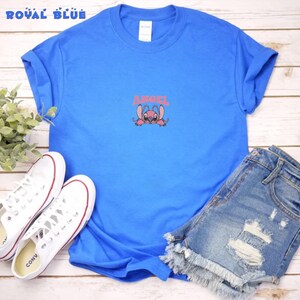 Pode incluir: Camiseta azul royal com um personagem de desenho animado rosa e a palavra "ANGEL" em rosa. A camiseta &eacute; combinada com shorts jeans e t&ecirc;nis brancos.