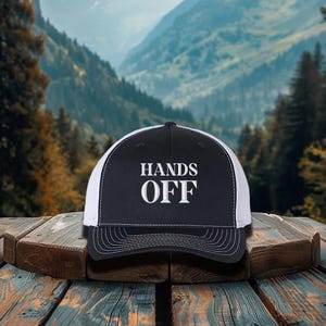 Pu&ograve; includere: Cappellino da camionista nero e bianco con la scritta "HANDS OFF" in lettere bianche. Il cappellino ha un pannello frontale nero e un retro in rete bianca. Il cappellino &egrave; appoggiato su una superficie di legno con uno sfondo montuoso.