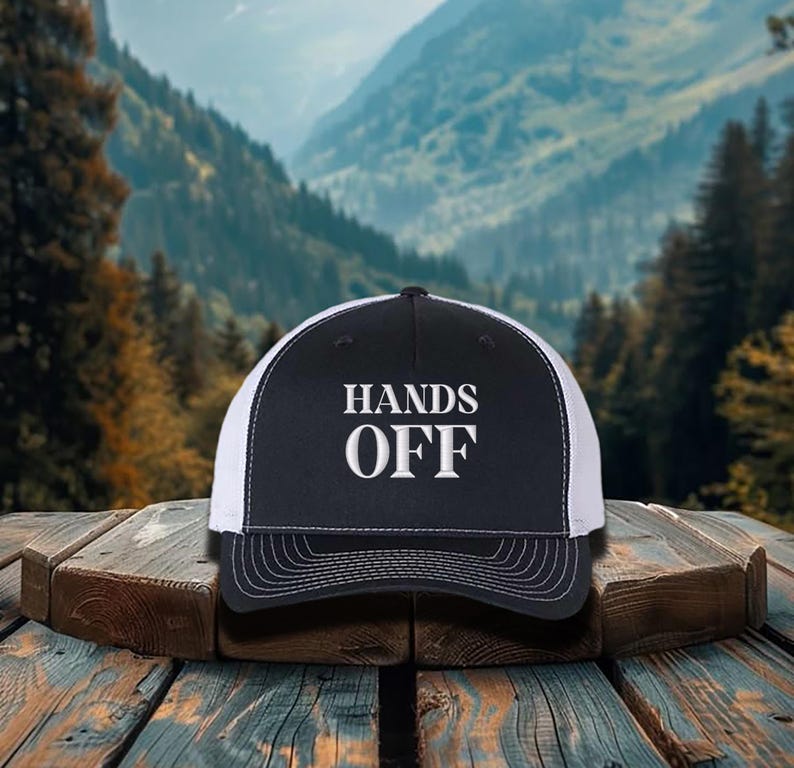 Pu&ograve; includere: Cappellino da camionista nero e bianco con la scritta "HANDS OFF" in lettere bianche. Il cappellino ha un pannello frontale nero e un retro in rete bianca. Il cappellino &egrave; appoggiato su una superficie di legno con uno sfondo montuoso.