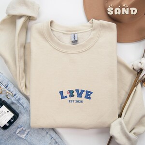 May include: A cream-coloured sweatshirt with the word "LOVE" embroidered in blue, featuring a cartoon character design. The text "EST 2026" is below the word "LOVE". A brown hat with the word "SAND" is in the background.