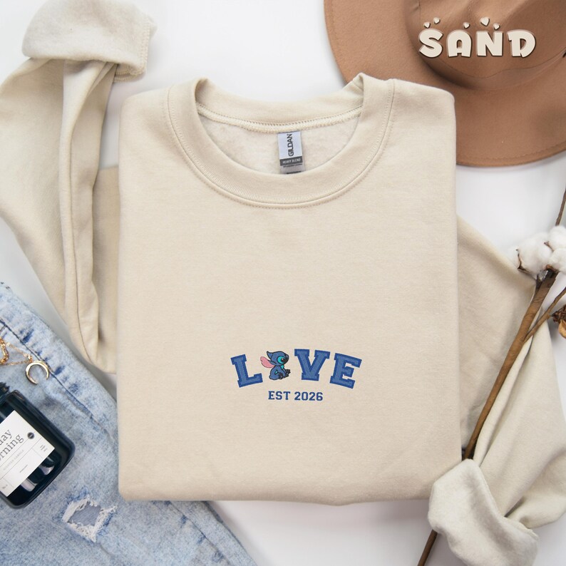 May include: A cream-coloured sweatshirt with the word "LOVE" embroidered in blue, featuring a cartoon character design. The text "EST 2026" is below the word "LOVE". A brown hat with the word "SAND" is in the background.