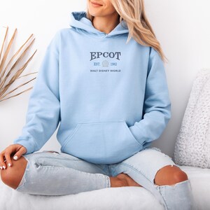 May include: A light blue hoodie with the text "EPCOT EST. 1982 WALT DISNEY WORLD" printed in navy blue. The hoodie has a front pocket and a hood with a drawstring. The person is wearing light blue jeans with ripped knees.