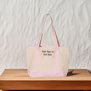 May include: A beige canvas tote bag with light pink trim and handles. The bag features the text "Your logo or text here" on the front pocket. A simple, practical design.
