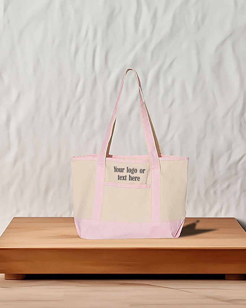May include: A beige canvas tote bag with light pink trim and handles. The bag features the text "Your logo or text here" on the front pocket. A simple, practical design.