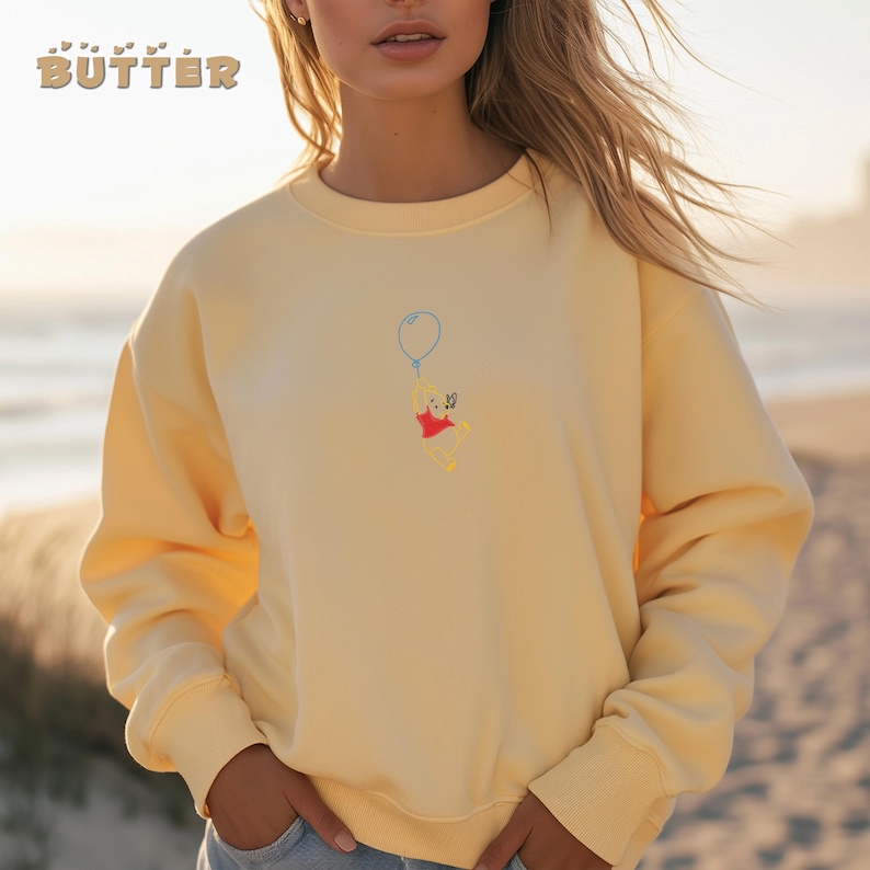 May include: A yellow sweatshirt with a Winnie the Pooh graphic. Pooh is holding a blue balloon and is flying in the air. The graphic is embroidered on the sweatshirt.