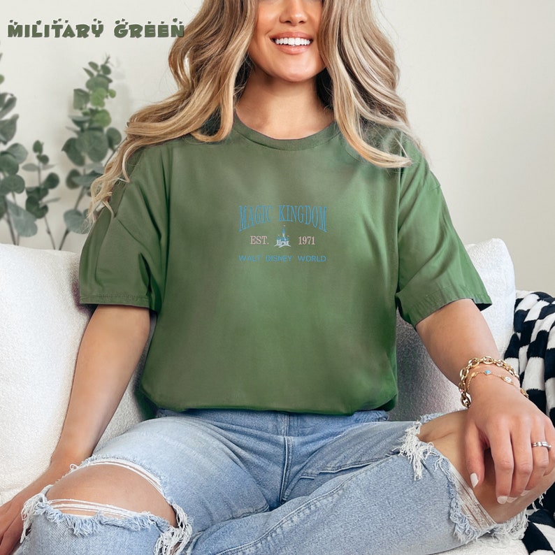 May include: A woman wearing a green t-shirt with the text "Magic Kingdom Est. 1971 Walt Disney World" in blue.