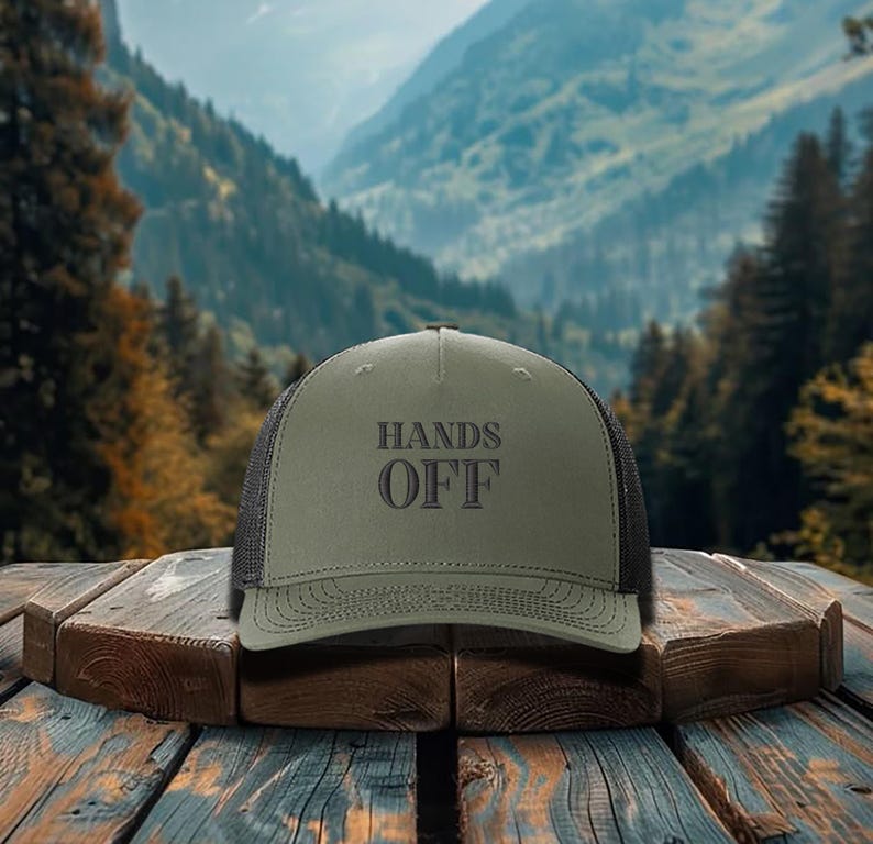 Pu&ograve; includere: Cappellino da camionista verde oliva con la scritta "HANDS OFF" in grigio scuro. Il cappello ha un retro in rete e una visiera curva. Il cappello &egrave; appoggiato su una superficie di legno con uno sfondo montuoso.
