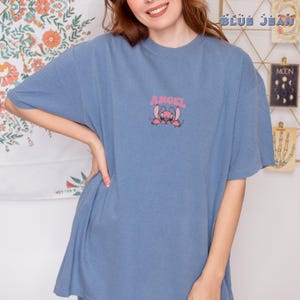 Puede incluir: Una camiseta azul con un gr&aacute;fico rosa de un personaje de dibujos animados y la palabra "ANGEL" encima. La camiseta es de color azul liso y tiene mangas cortas.
