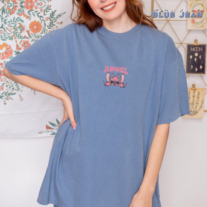 Puede incluir: Una camiseta azul con un gr&aacute;fico rosa de un personaje de dibujos animados y la palabra "ANGEL" encima. La camiseta es de color azul liso y tiene mangas cortas.