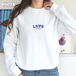 May include: White crew neck sweatshirt featuring the word "LOVE" in blue block letters, a floral design, and the text "EST 2026". The sweatshirt is made of a soft material. The word "white" is printed in the top left corner.
